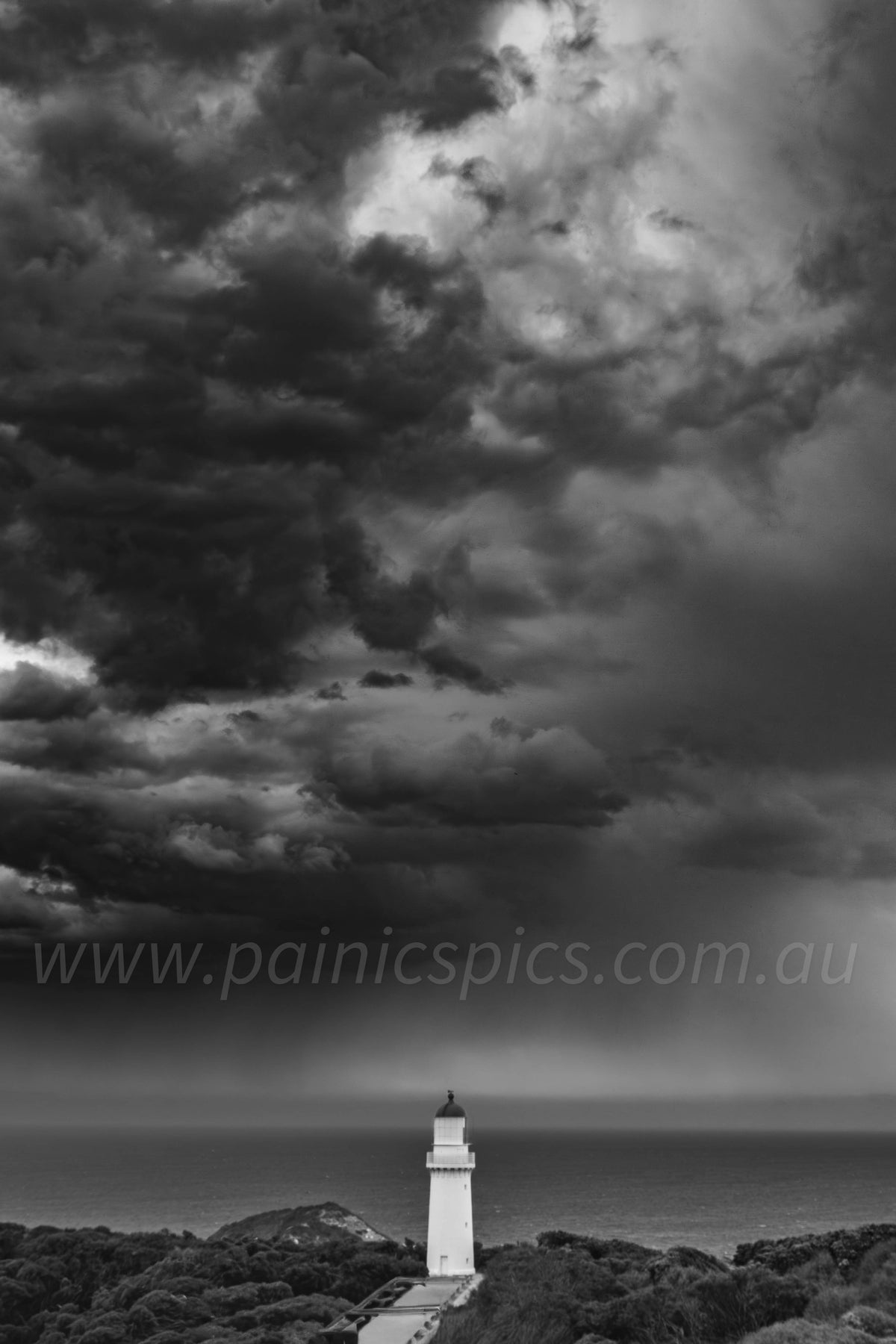 Black and white landscape photograph of a lighthouse situated on a rocky headland overlooking a calm sea under a dramatic, stormy sky with heavy clouds.