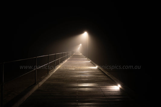 A long wooden boardwalk at night with glowing overhead and ground lights, fading into a misty darkness in the distance.