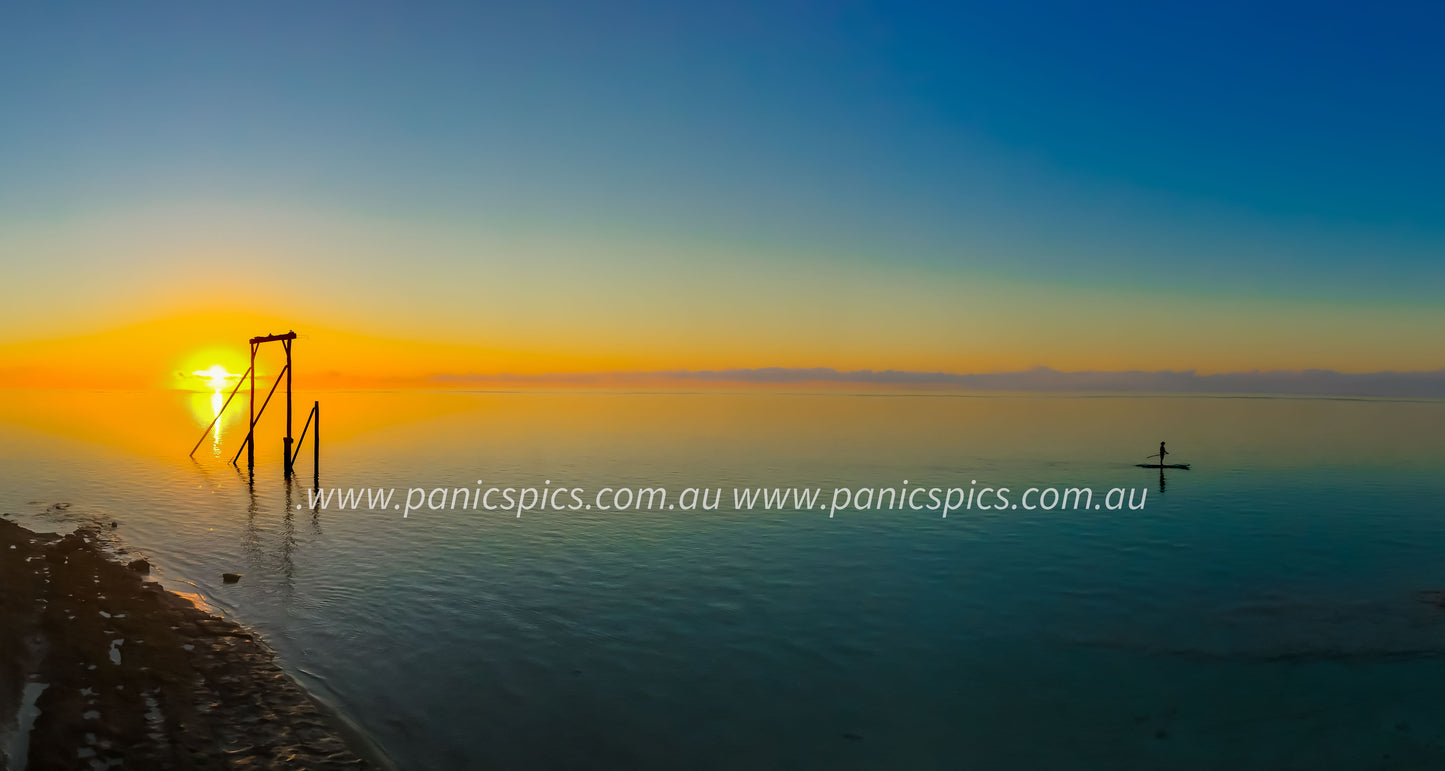 Sunset over a calm body of water with a wooden structure and a person on a surfboard.