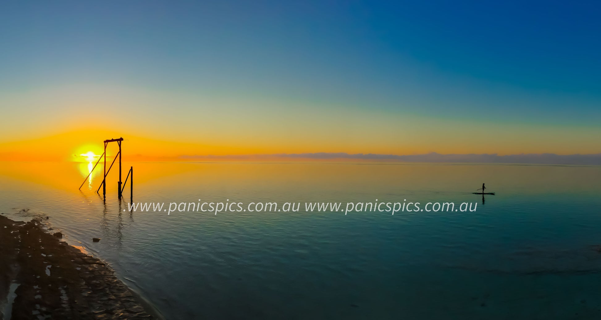 Sunset over a calm body of water with a wooden structure and a person on a surfboard.